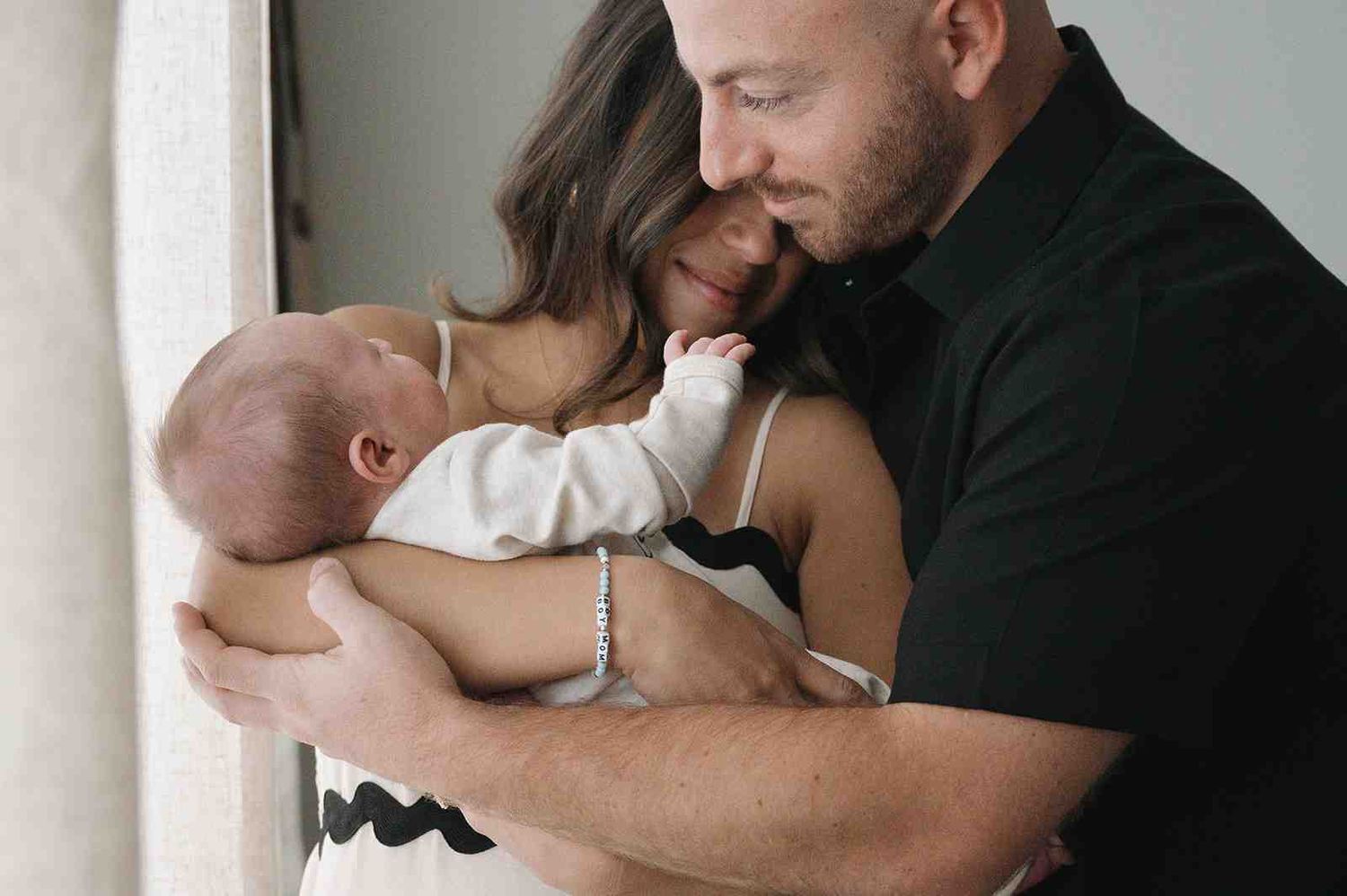 Parents share tender moment while holding their newborn baby next to a bright window.