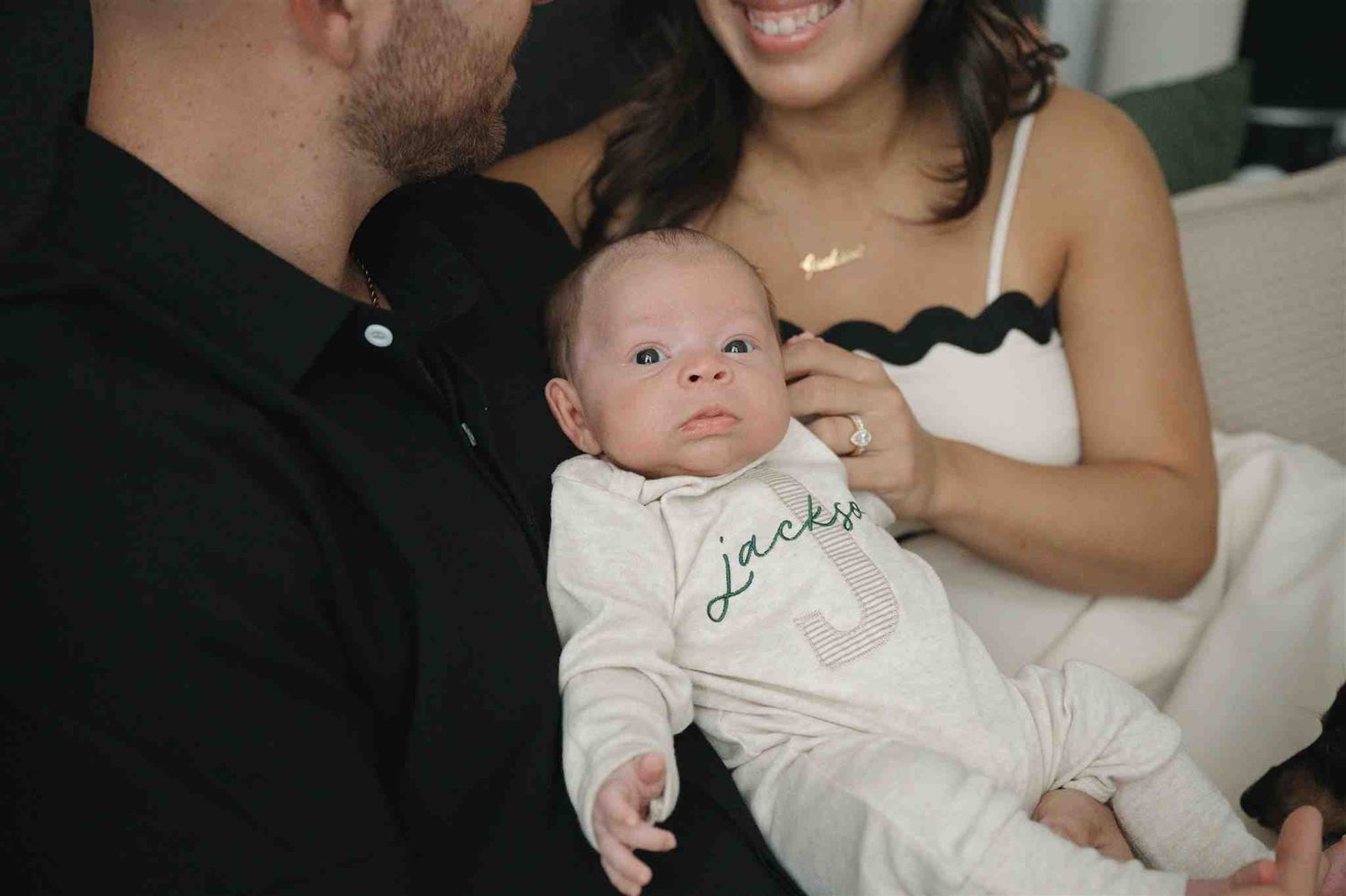 A baby wearing white clothing with 'Jackson' embroidered sits between two adults on a couch at home.