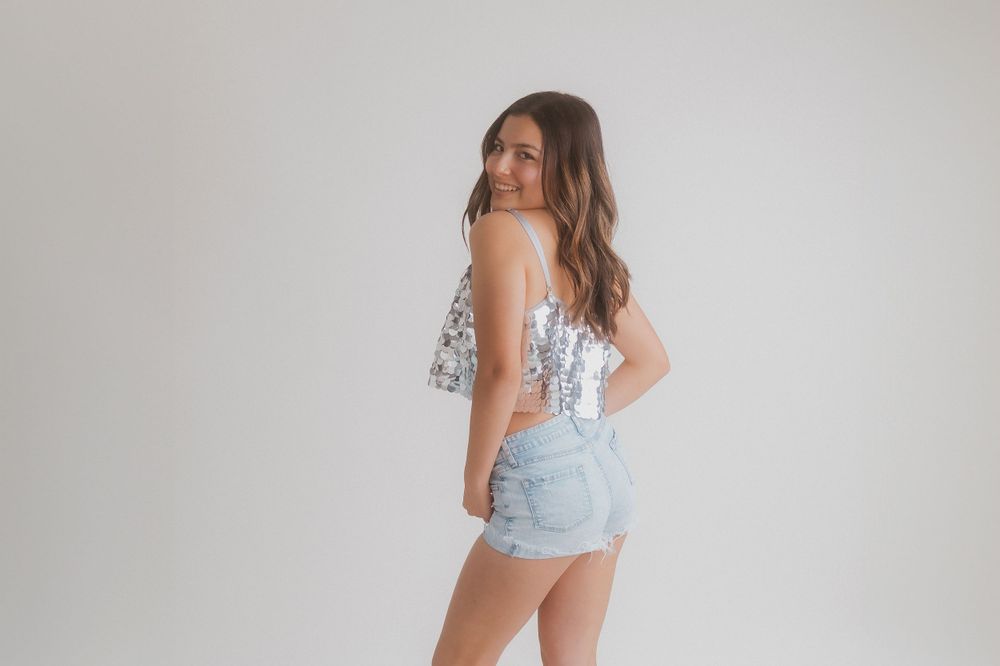 Portrait from behind showing sequin top and denim shorts against neutral background.
