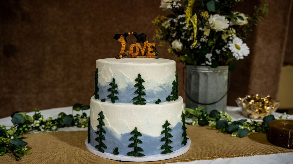 A two-tier white wedding cake decorated with silhouettes of pine trees and a rustic LOVE topper.