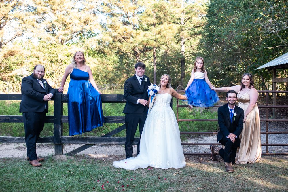 A wedding party poses together outdoors by a wooden fence on an autumn day wearing formal attire.