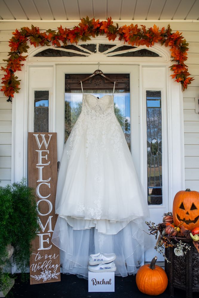 A wedding dress hangs on a front porch decorated with fall leaves, pumpkins, and a welcome sign for an autumn ceremony.