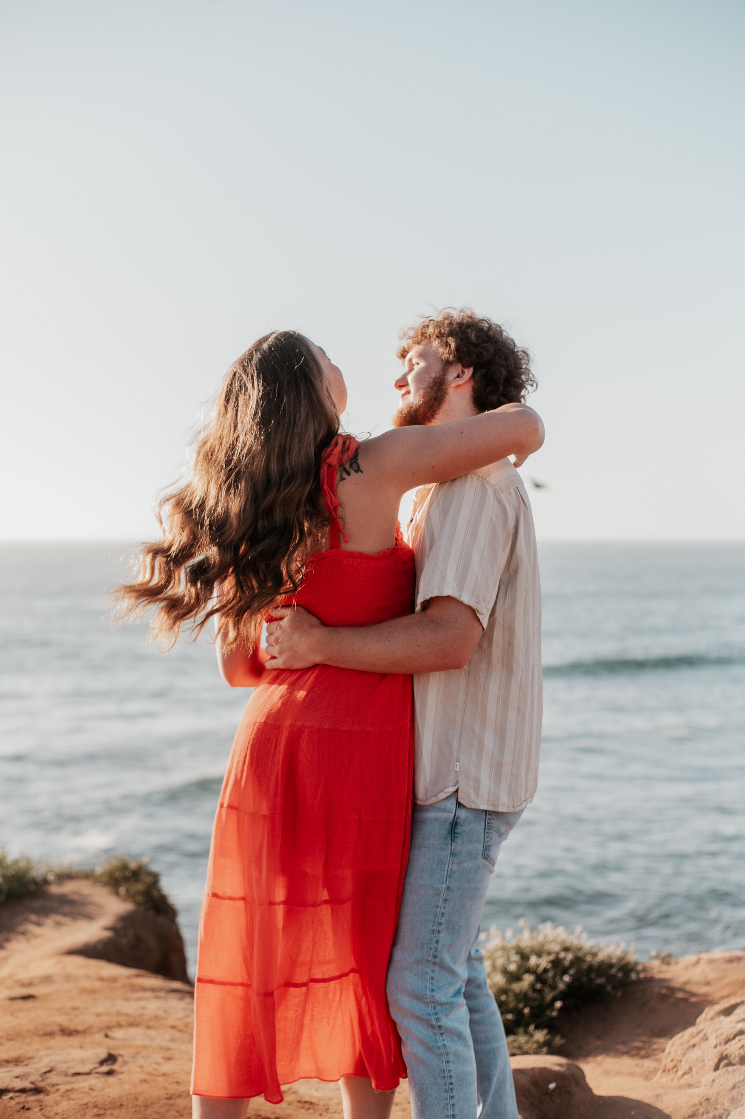 Romantic couple embraces on a scenic coastal cliff during golden hour, with ocean waves in the background.