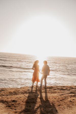 Sepia-toned silhouettes of couple standing on beach during sunset with long shadows.