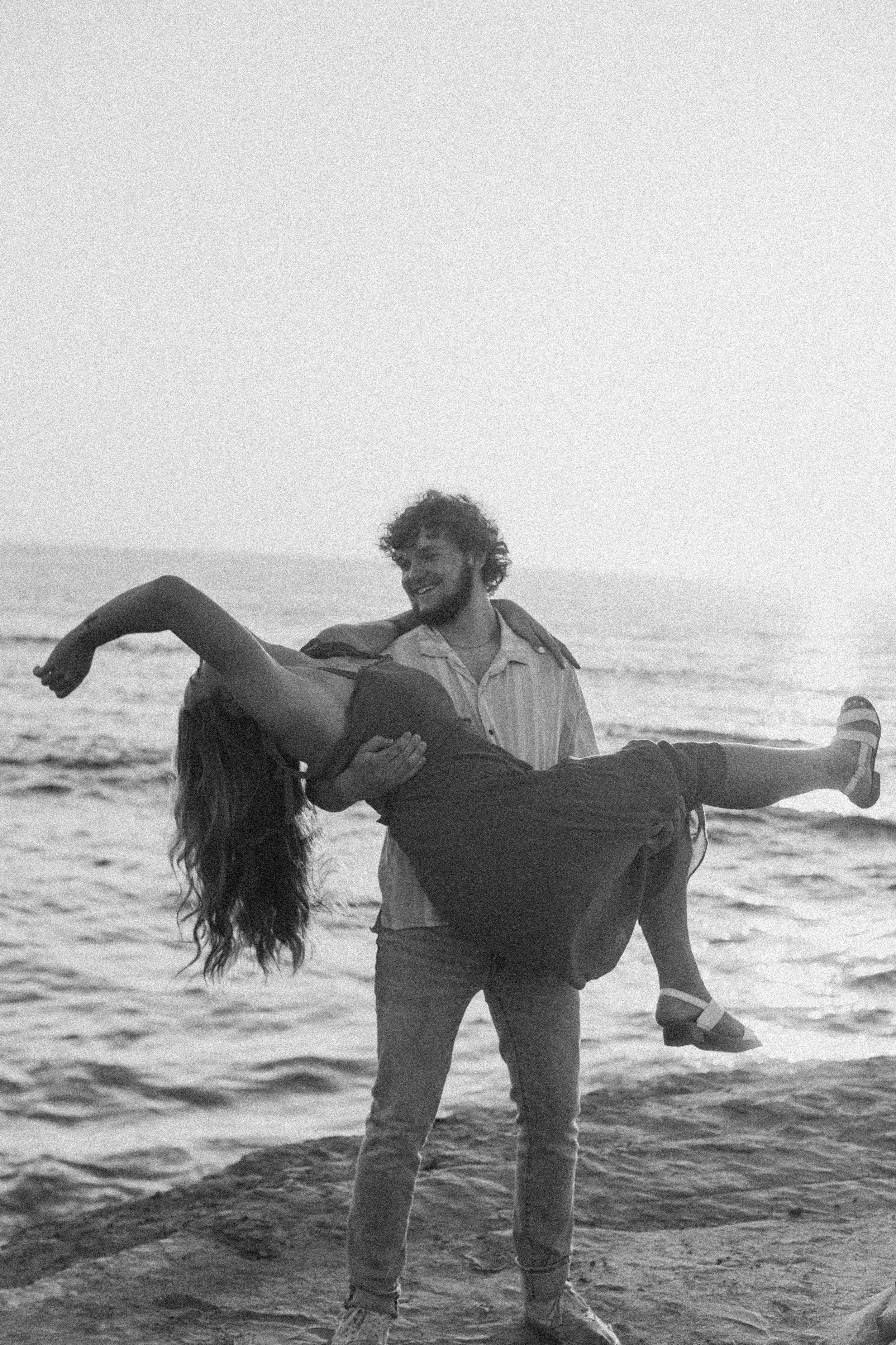 Romantic couple poses together in black and white photos on a beach with ocean waves in background.