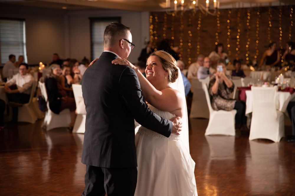 A couple shares their first dance at a wedding reception with string lights and white decorated tables in the background.