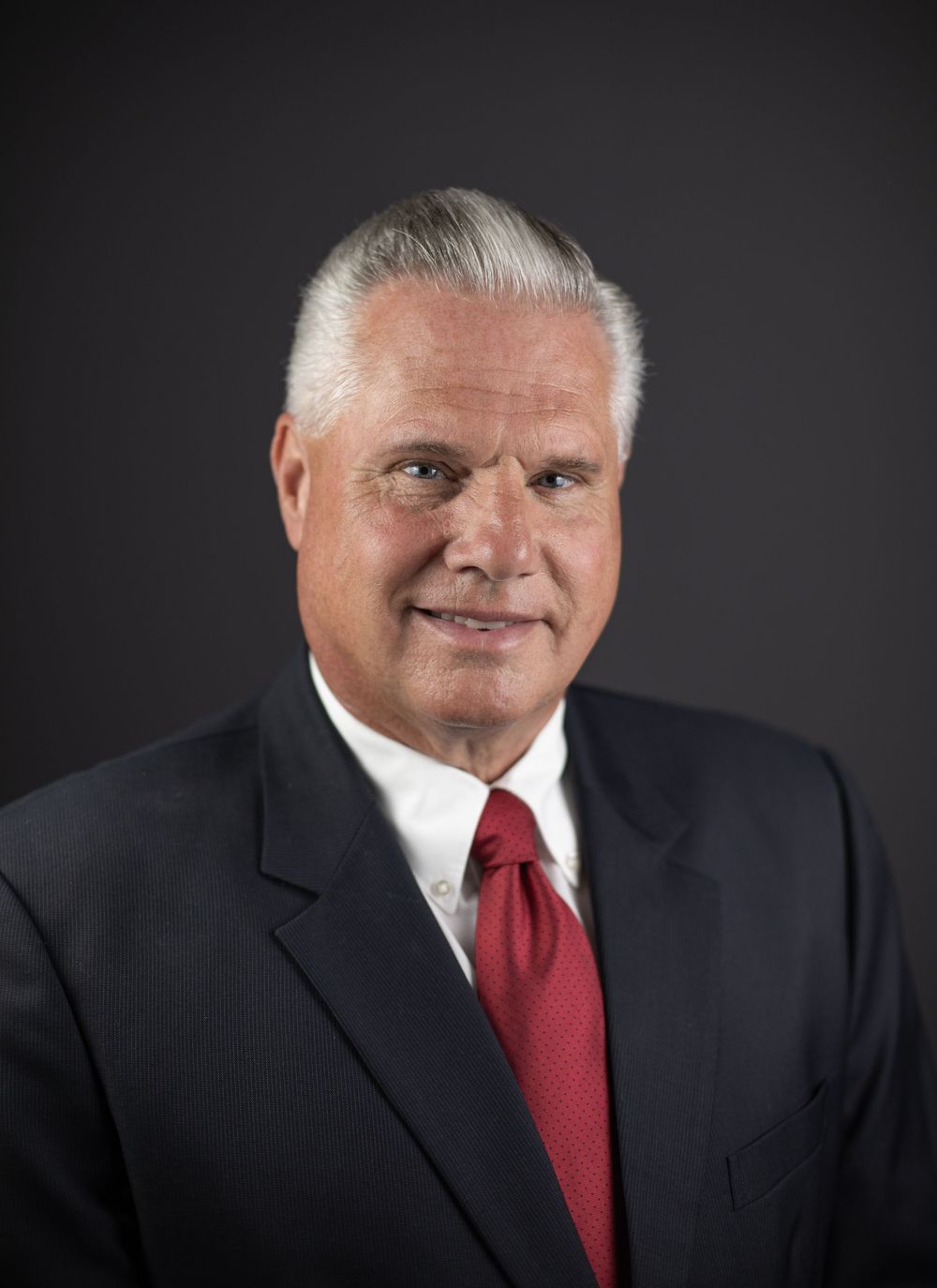 Professional headshot of a senior man with white hair wearing a dark suit and red tie against a gray background.