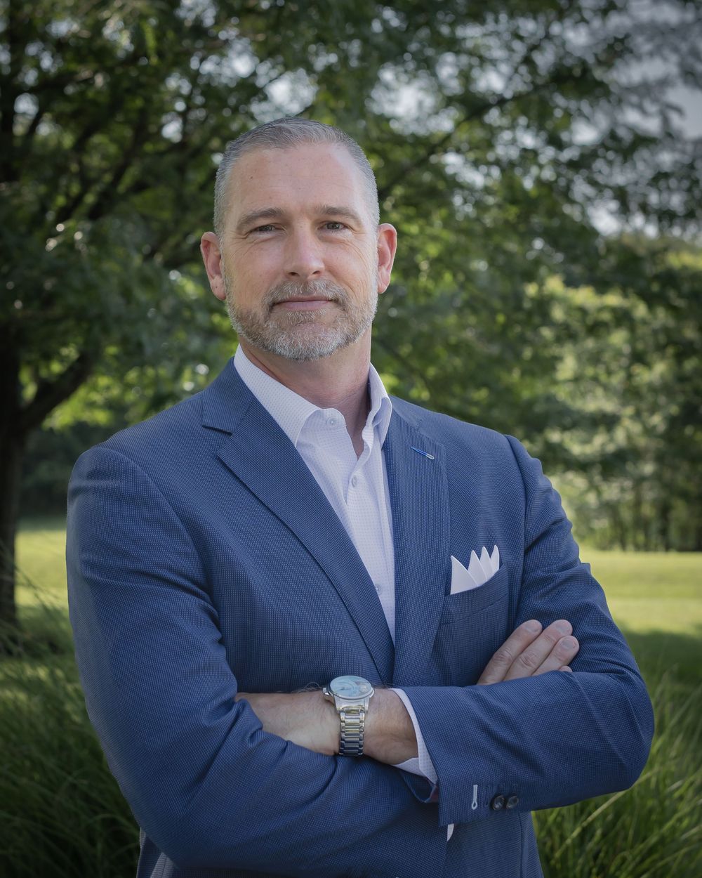 Professional man in blue blazer with arms crossed poses outdoors among green trees for a business portrait.