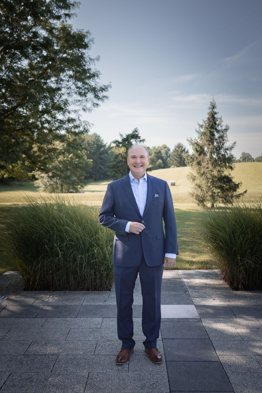 Professional man in navy suit smiling outdoors on stone path with lush green trees and golf course behind him.