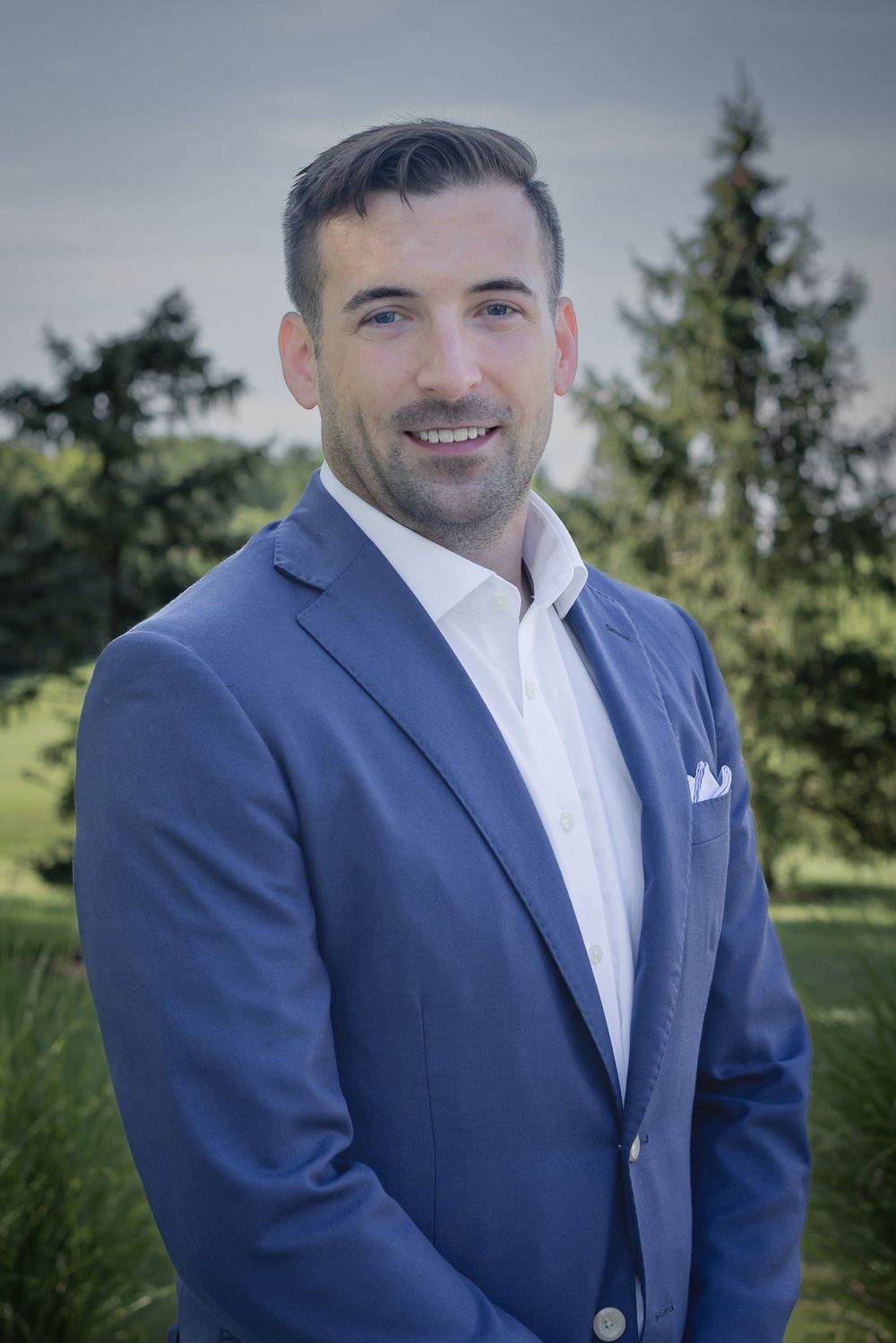 A smiling man in a blue blazer and white shirt poses outdoors with green trees in the background.