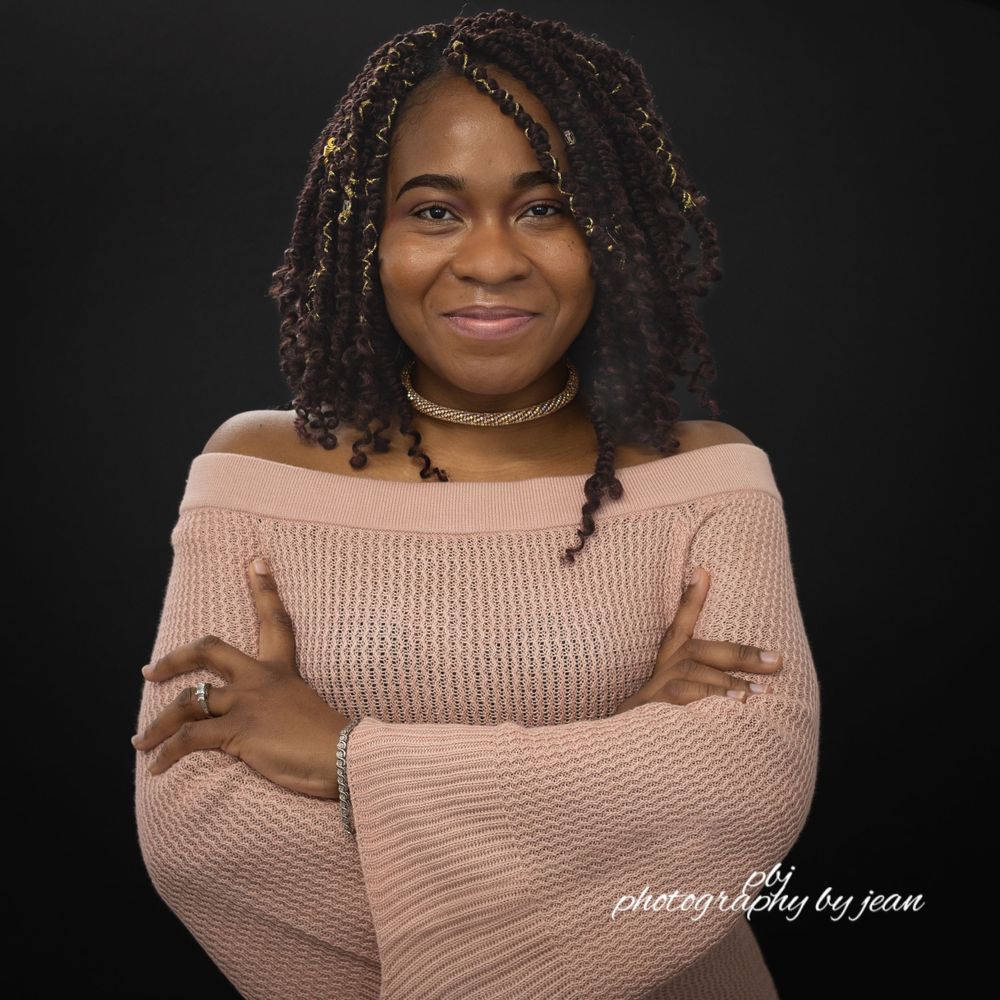 A person wearing a pink knit sweater poses against a dark background with curly hair and jewelry.