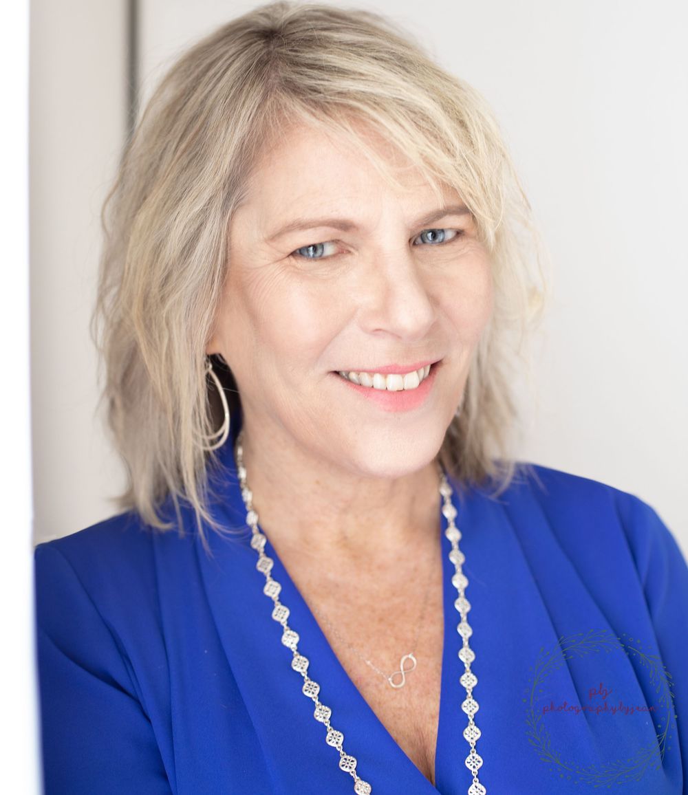 Smiling middle-aged woman with blonde hair wearing a blue blazer and white beaded necklace with hoop earrings.