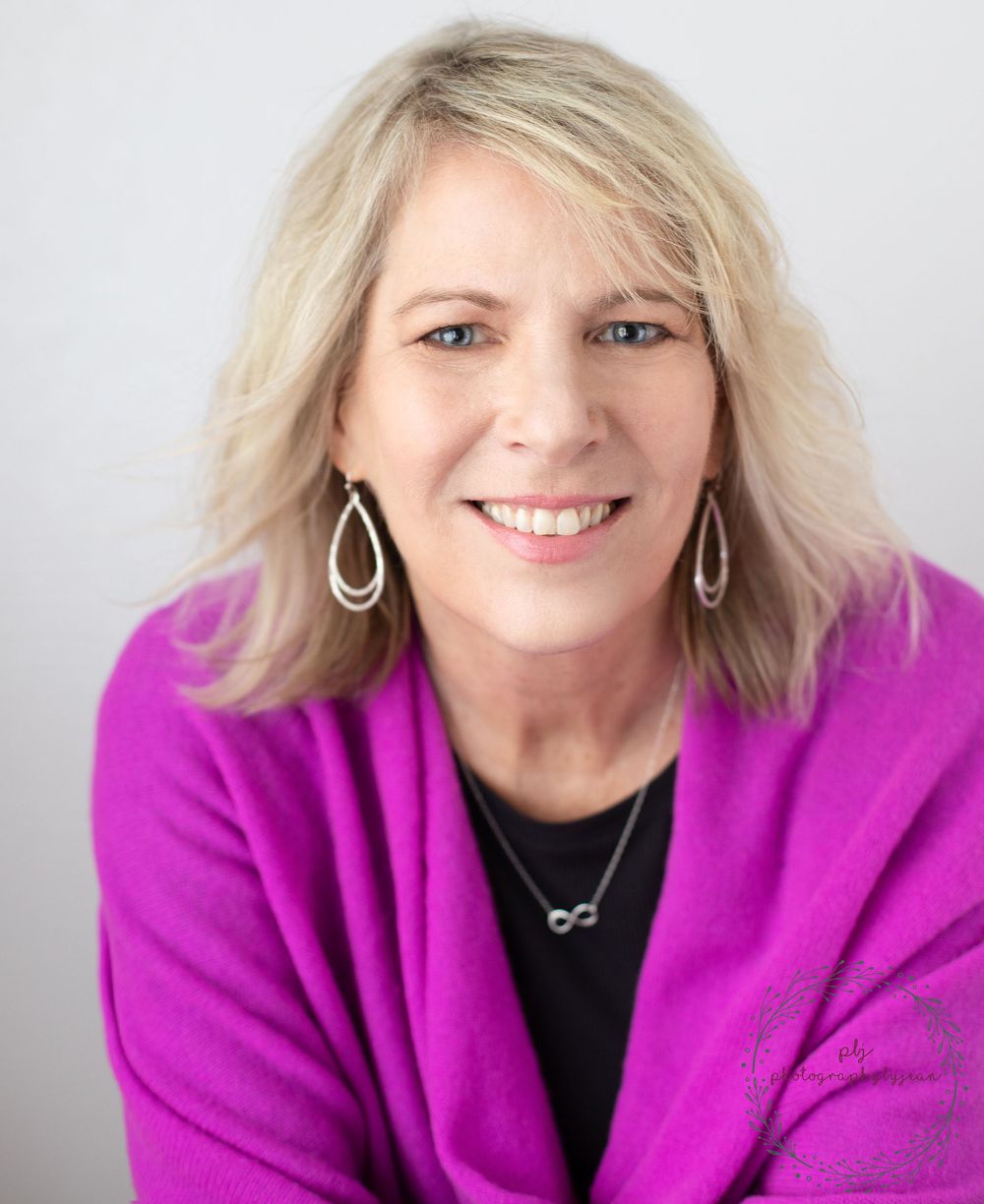 Smiling blonde woman in a bright magenta blazer and teardrop earrings poses for a professional headshot.