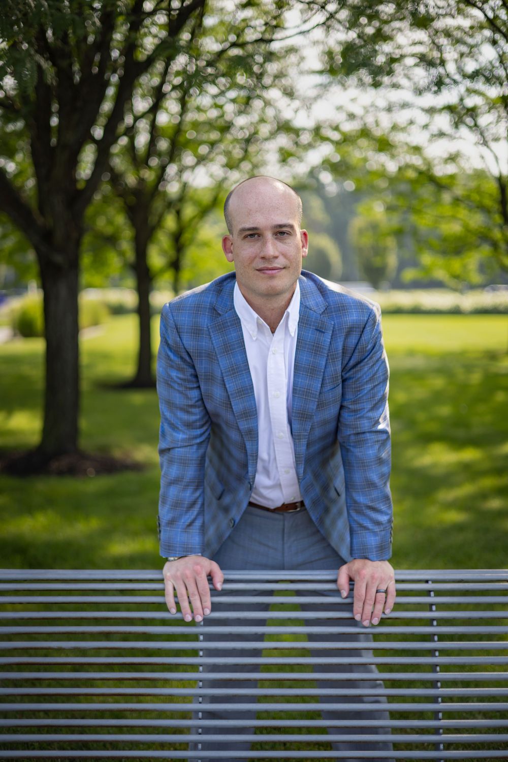 A bald man in a blue plaid blazer leans on a park bench, smiling outdoors among green trees.