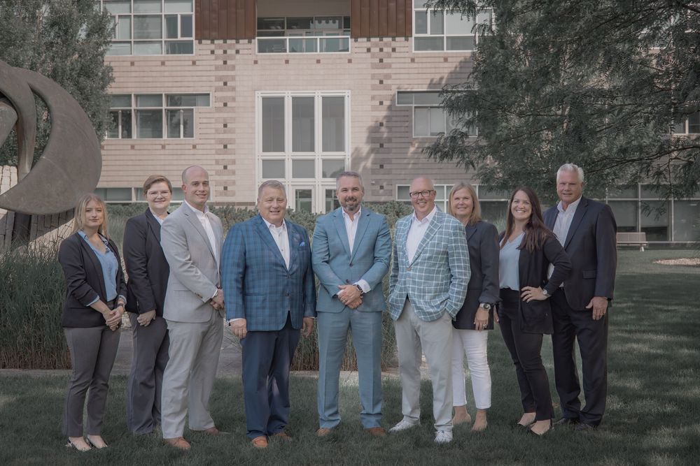 Group of eight business professionals posing outdoors in front of a modern brick building on a sunny day.