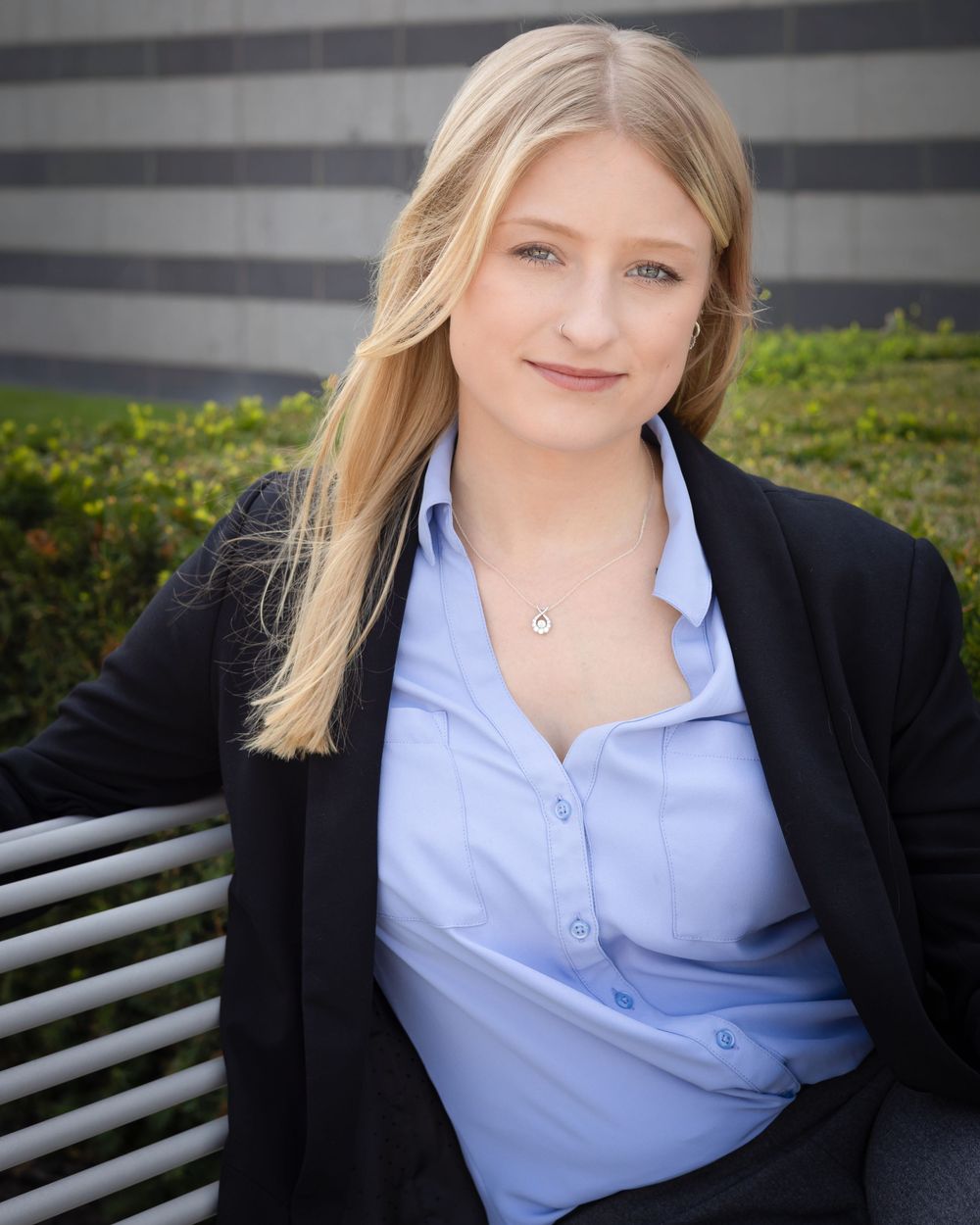 Young blonde woman in blue shirt and black blazer sitting on a bench outdoors for a professional headshot.