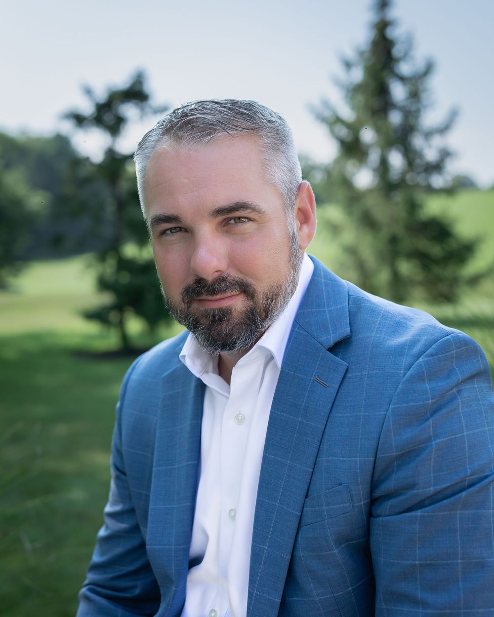 Professional man in blue blazer with gray hair and beard poses outdoors near green trees and golf course.