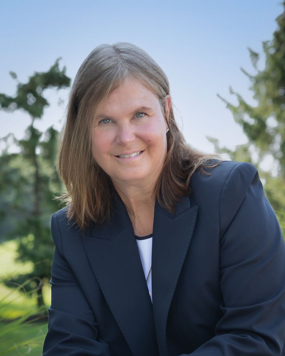Professional woman in navy blazer smiling outdoors with green trees in background, corporate headshot photo.