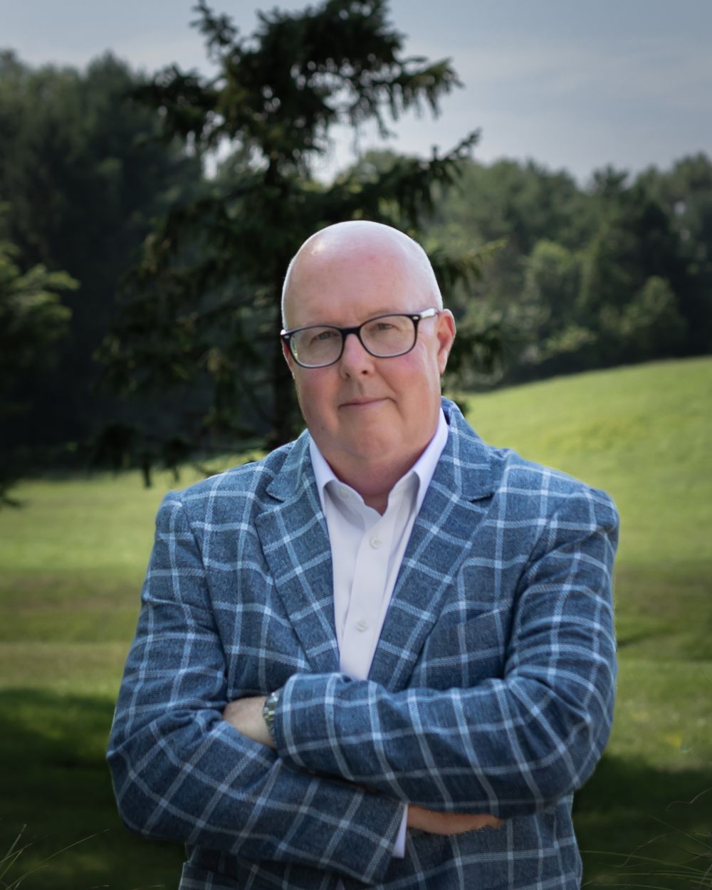 Professional man in plaid blazer with crossed arms standing outdoors on green lawn with trees.