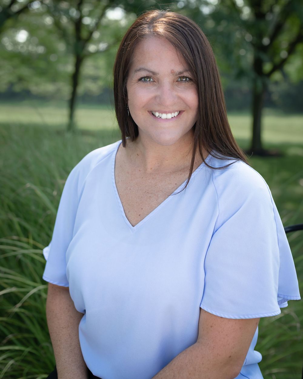 Smiling woman with brown hair wearing a light blue v-neck top outdoors with green trees in the background.