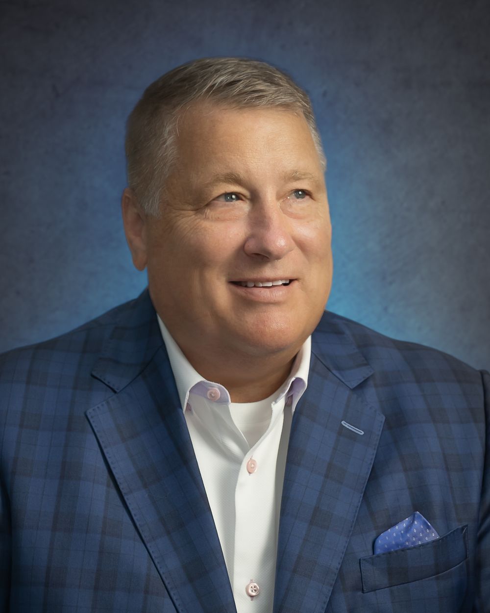 Professional headshot of a smiling middle-aged man in a blue plaid blazer and white shirt against a blue background.
