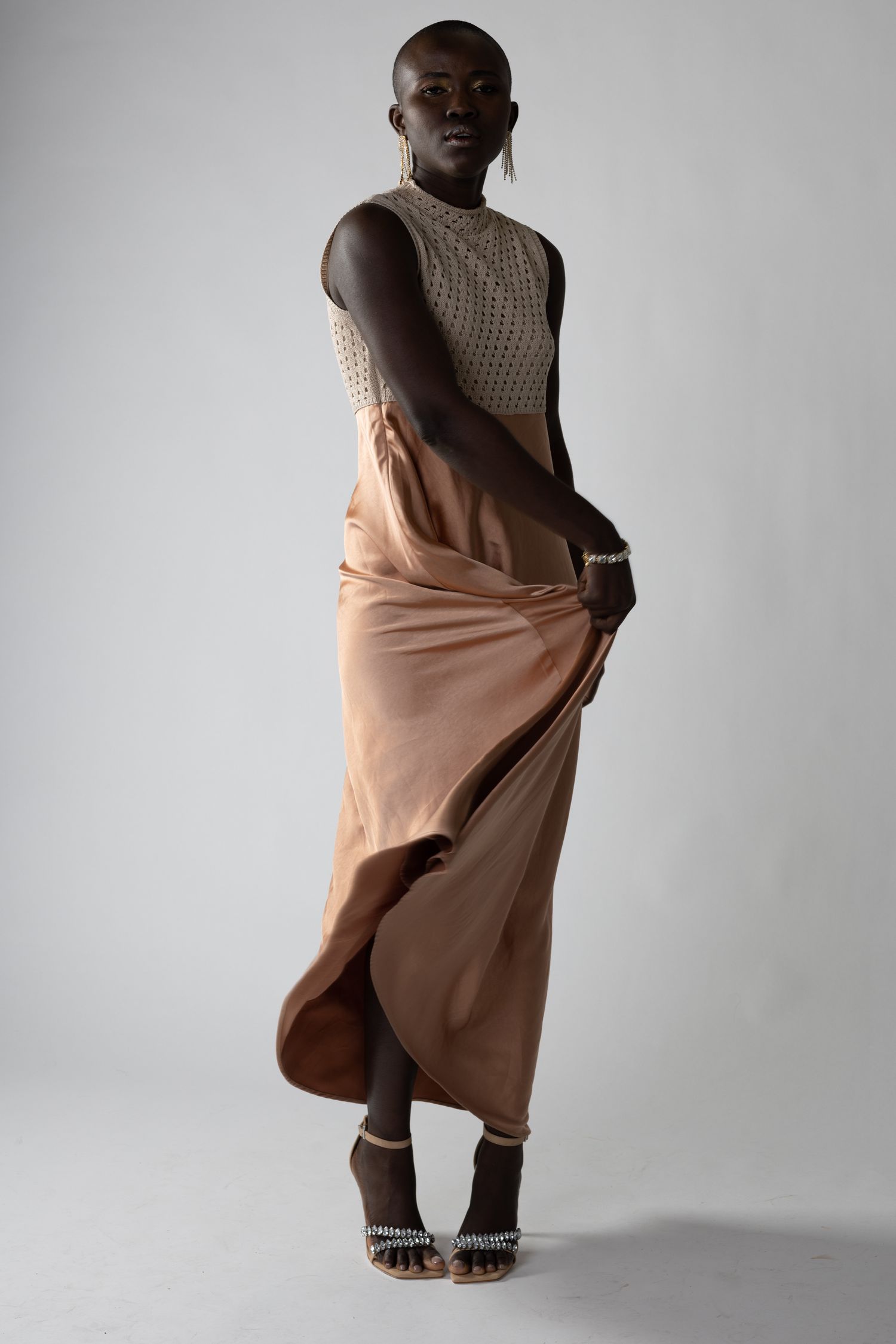 Fashion model in beige knit top and flowing skirt poses elegantly against grey background.