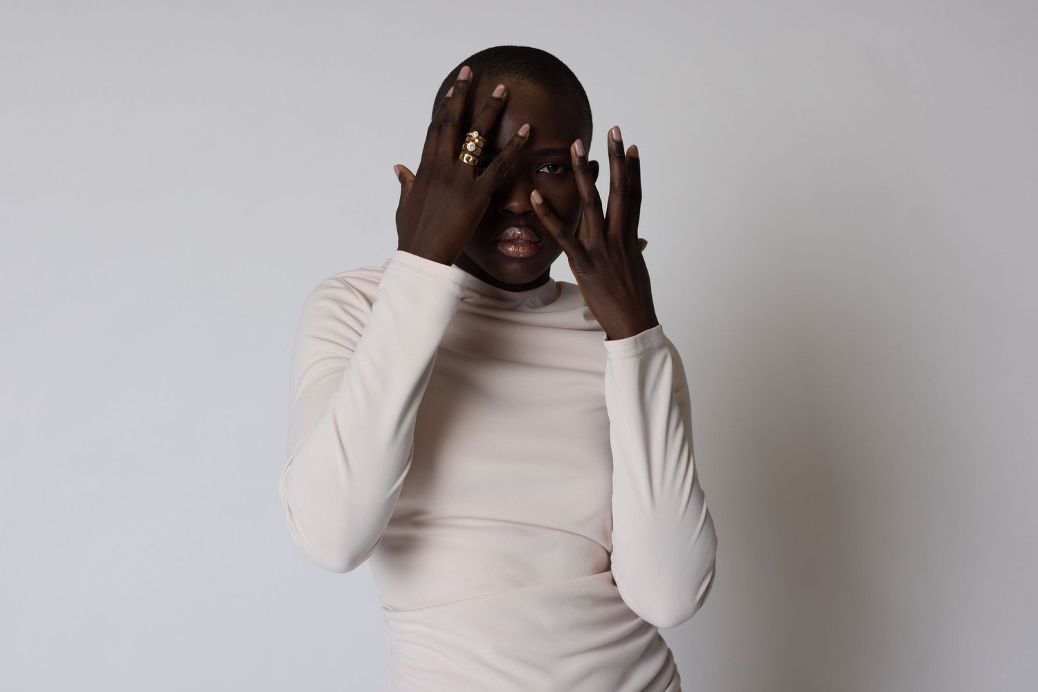 Artistic portrait in white turtleneck against grey background with dramatic hand gesture.