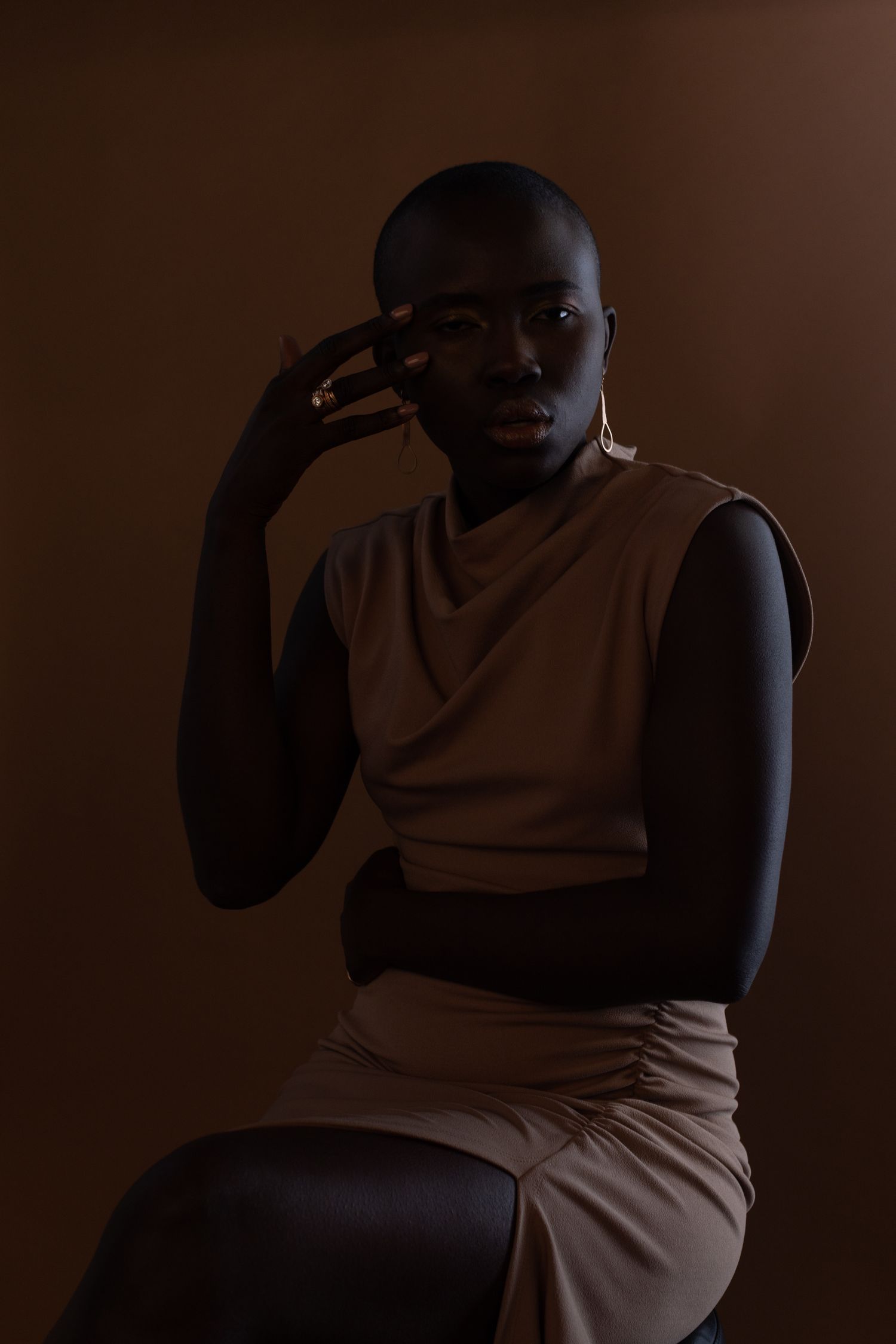 Artistic portrait with moody lighting showing figure in beige dress against dark brown backdrop.