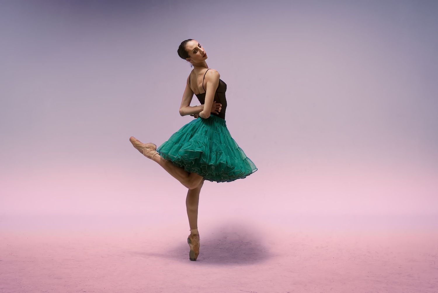 Ballet dancer in emerald green tutu performs graceful pose on pink background.