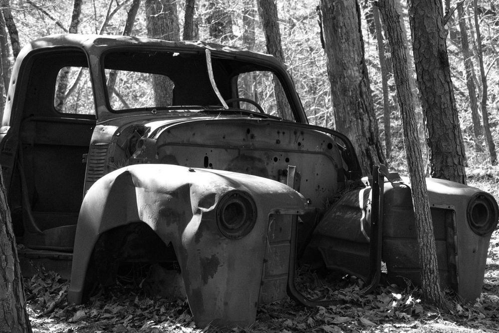 An abandoned vintage truck rusts among trees in a forest setting, photographed in black and white.
