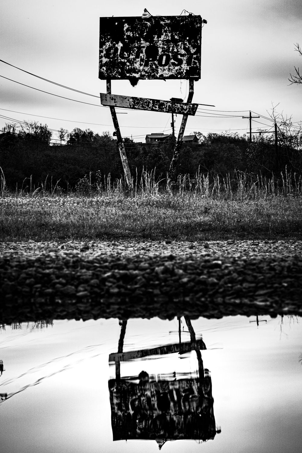 A weathered billboard sign reflects in water puddle at dusk, creating a mirror image.