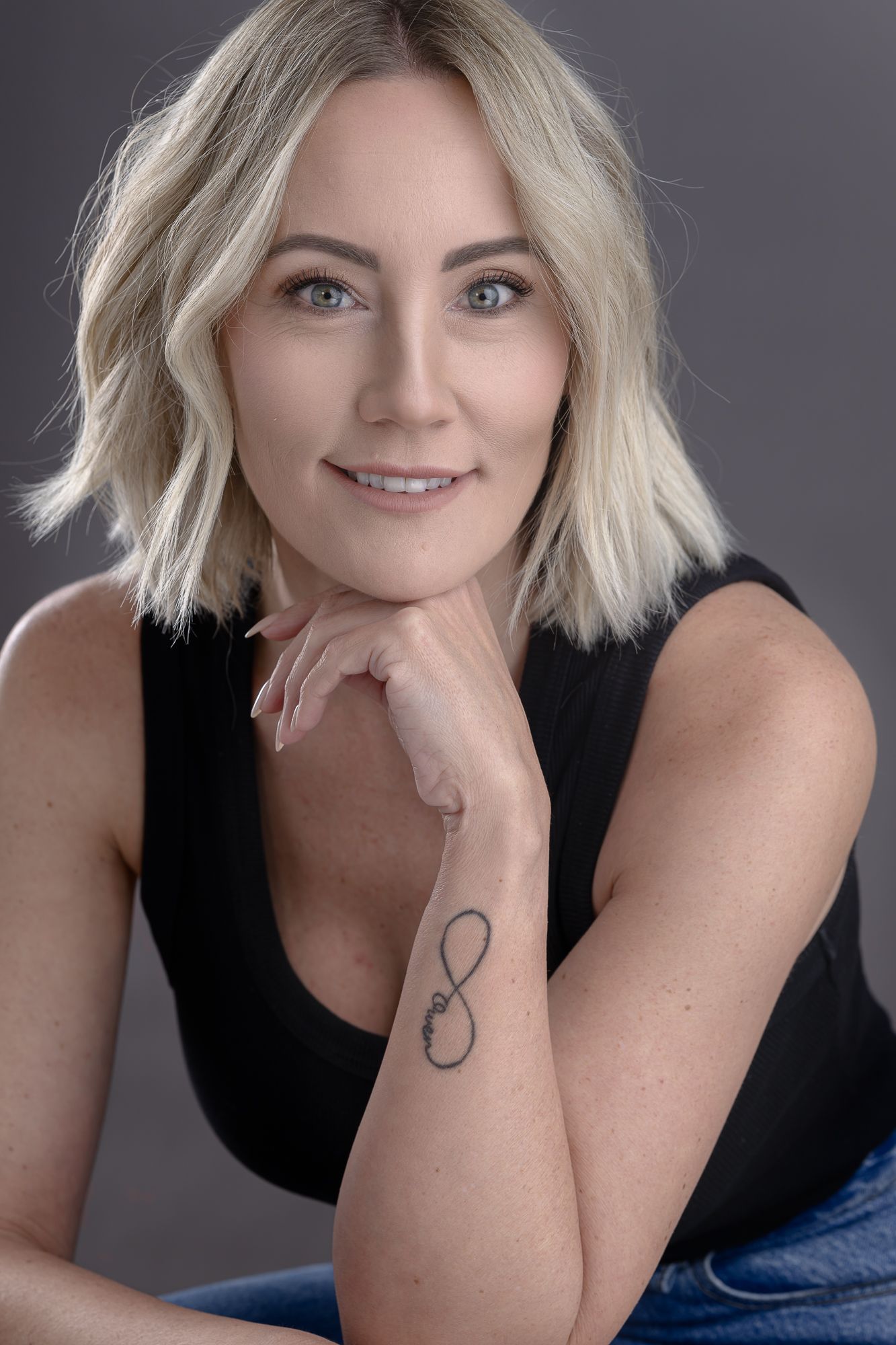 Portrait of woman with blonde wavy hair in black tank top showing infinity tattoo.