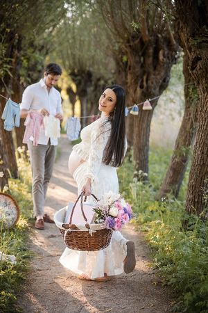 Romantic pregnant couple enjoys a picnic date under trees at sunset with flowers and basket on a forest path.