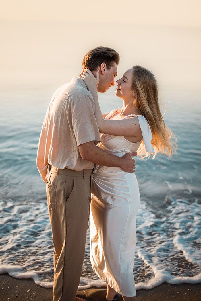 A romantic couple embraces on a beach at sunset with gentle waves washing up on the shore.