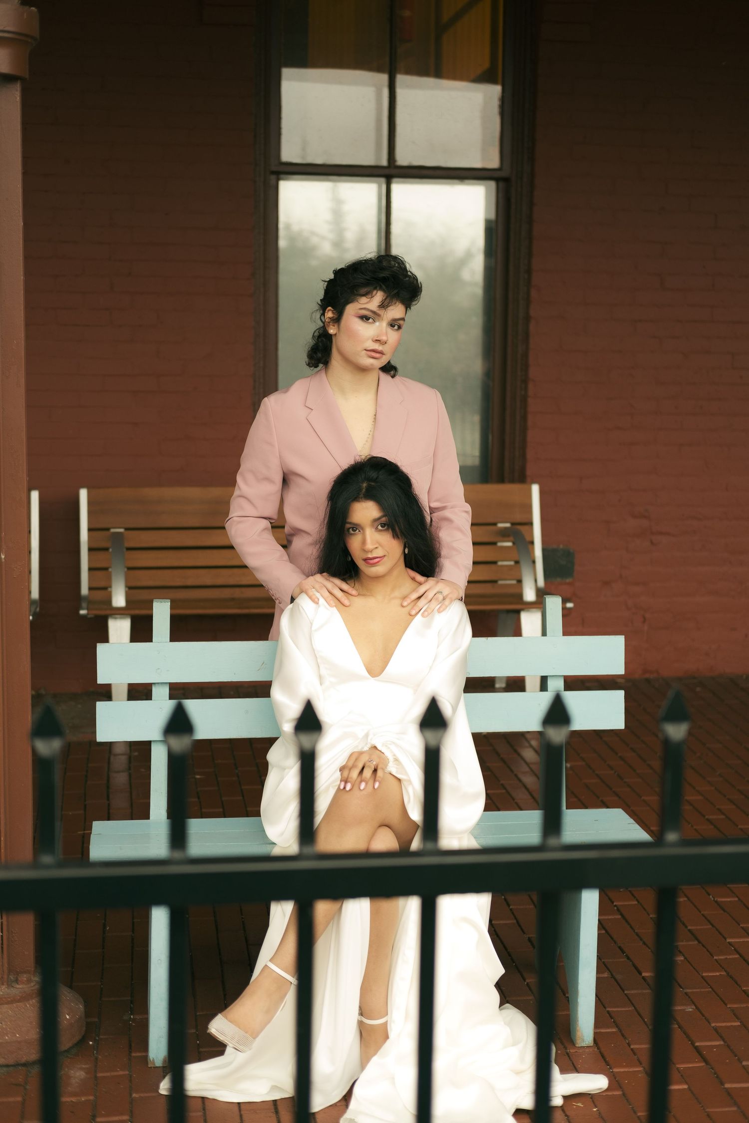 a young queer couple posing on a blue bench, one woman standing behind holding the shoulders of her bride as she sits