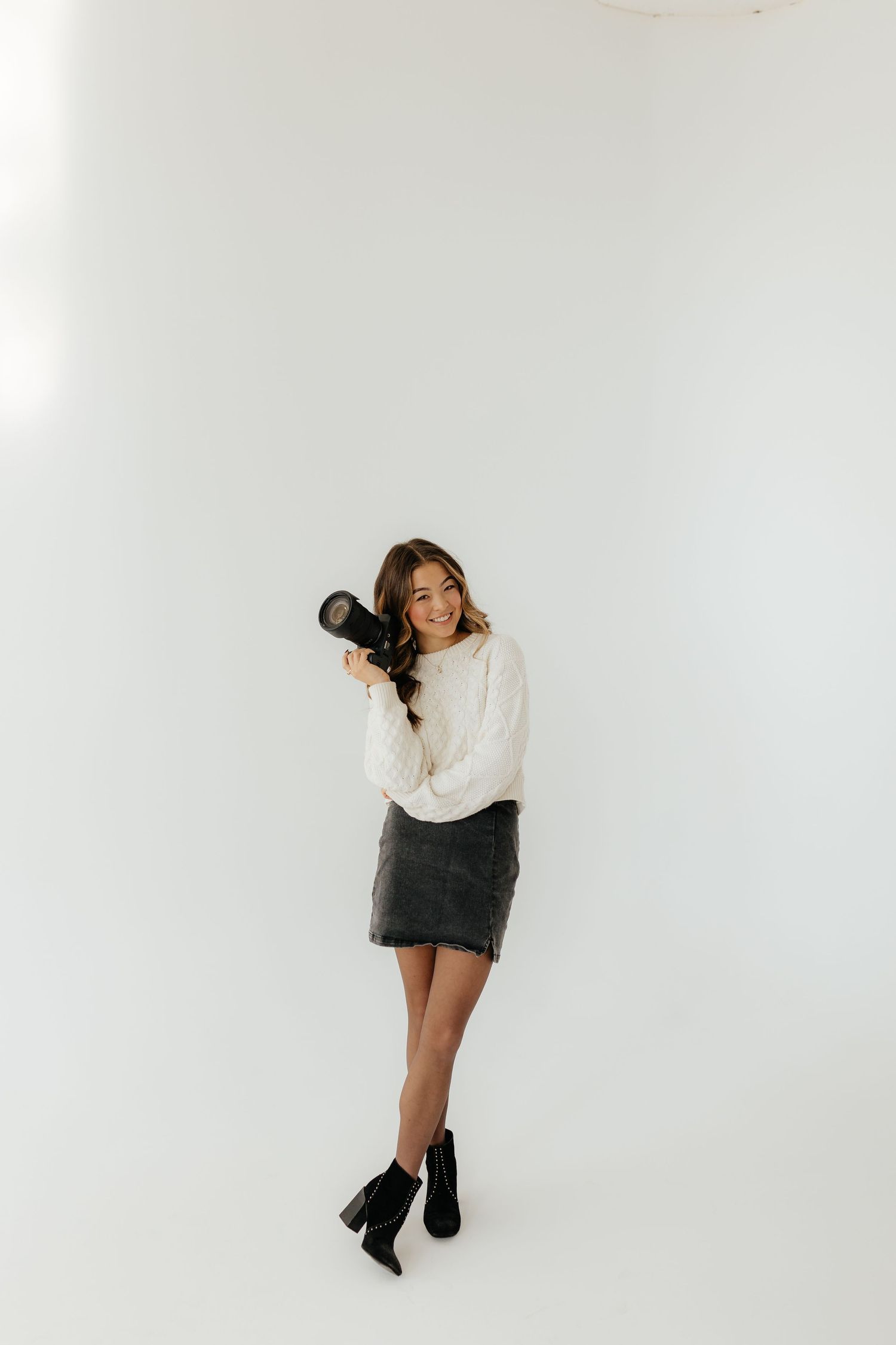 A smiling female photographer in a white sweater and dark skirt holds a camera in a bright white studio.