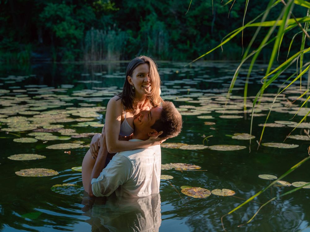 Couple embracing romantically in a serene lily pad lake surrounded by lush green trees in warm golden light.