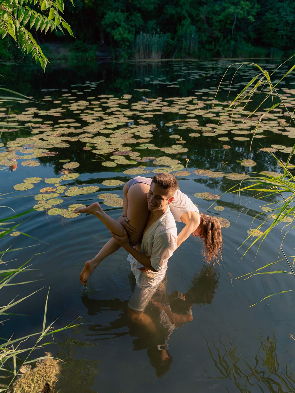 Two people sharing a romantic embrace while standing in a serene lily pad pond surrounded by lush greenery.