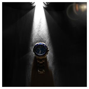 A wristwatch with blue face illuminated by dramatic spotlight beam in dark setting creating striking contrast.