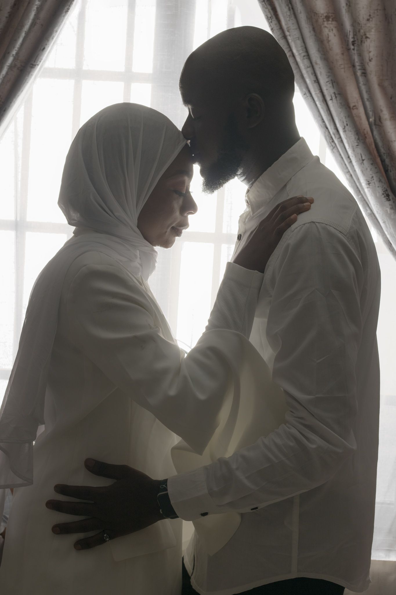 Intimate silhouette of couple in white against window light and curtains.