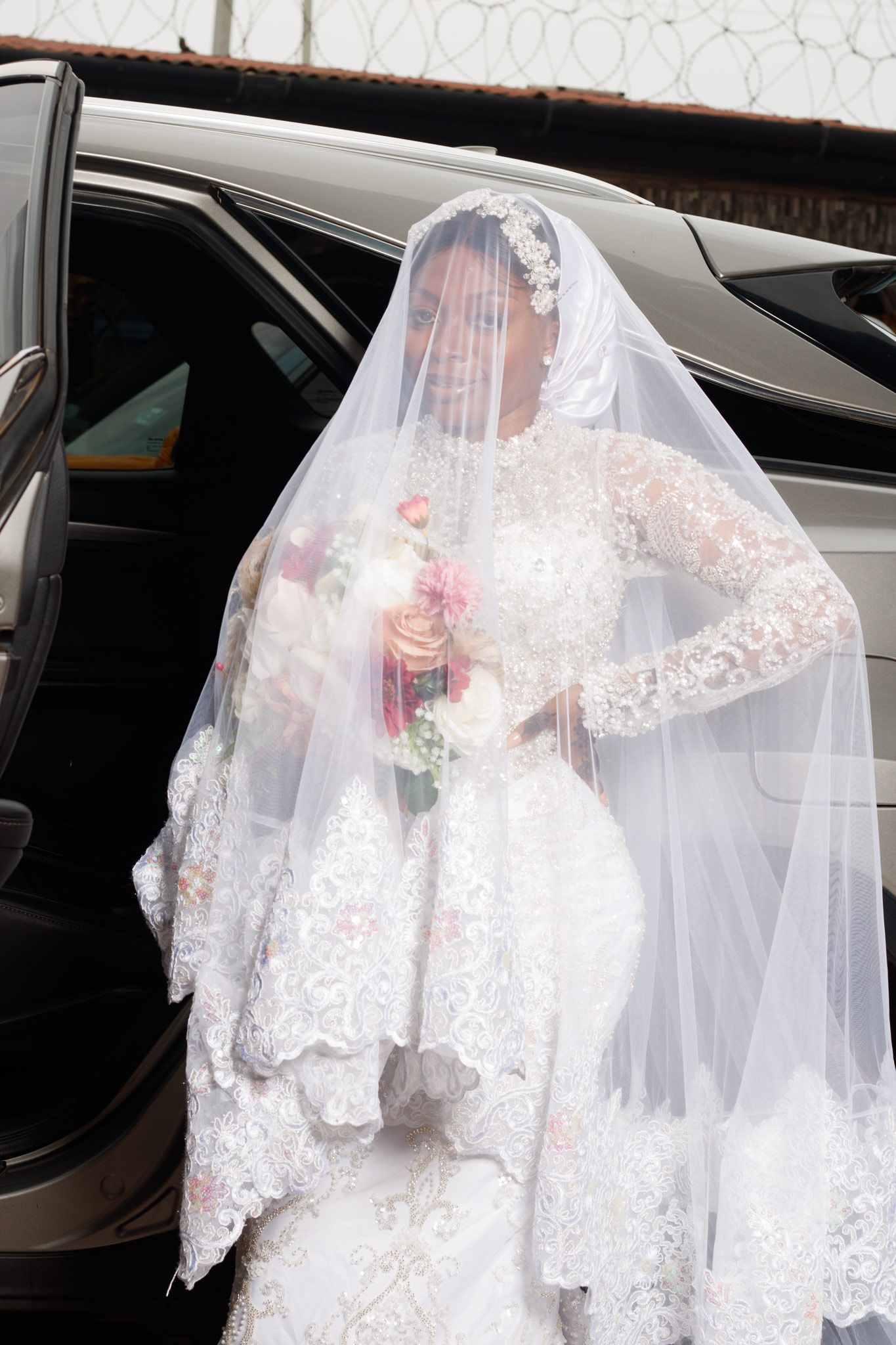 Elegant bride in a white lace long-sleeve wedding gown with detailed veil stepping out of a luxury car.