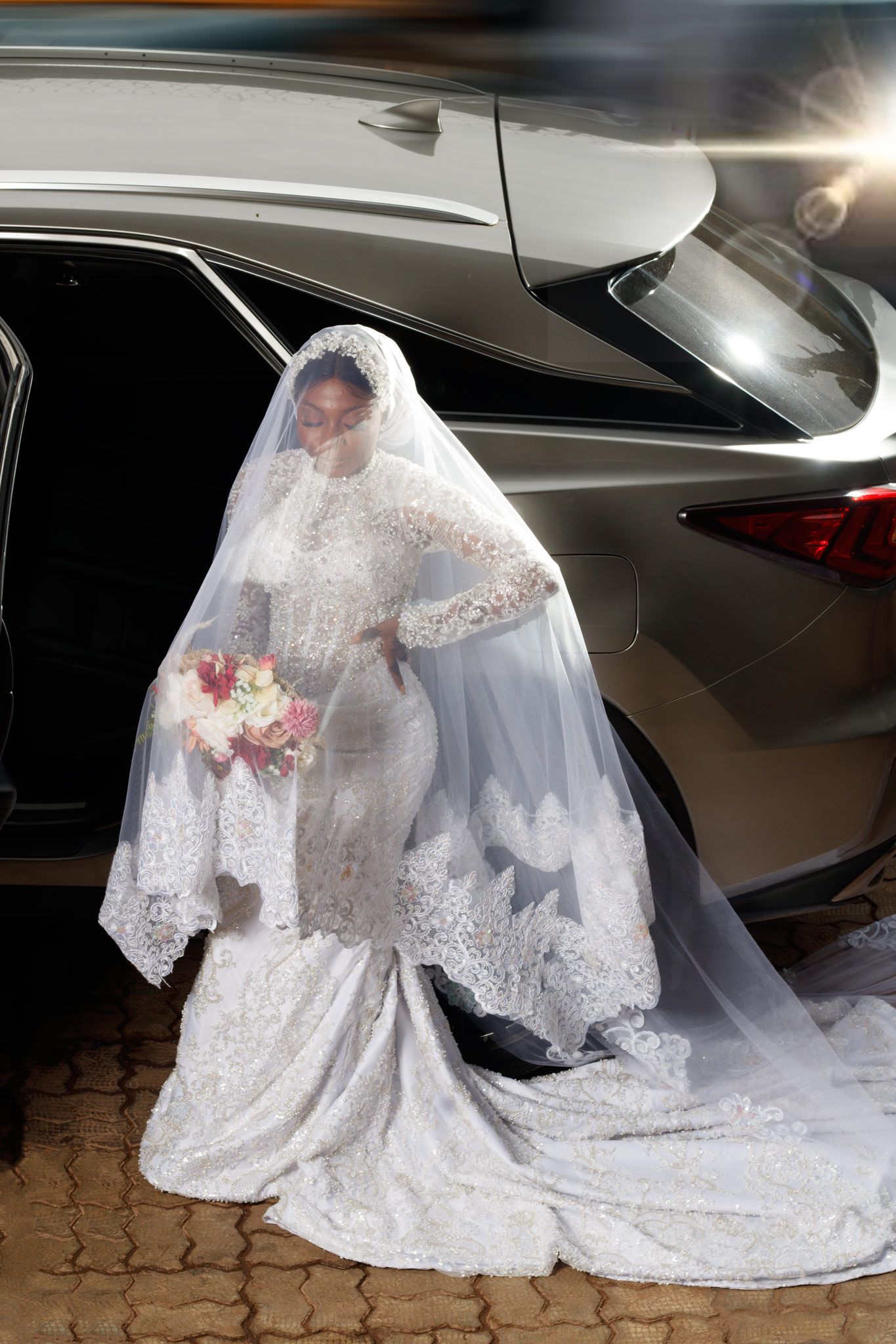 A stunning bridal gown with lace details and flowing train shown against a silver car exterior.