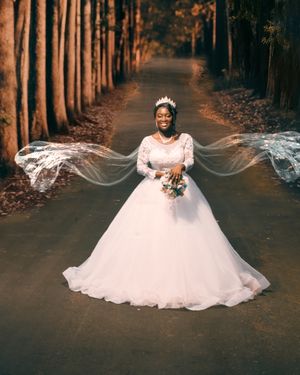 A bride in a stunning white ballgown and crown stands on a tree-lined path as her veil flows dramatically in the breeze.