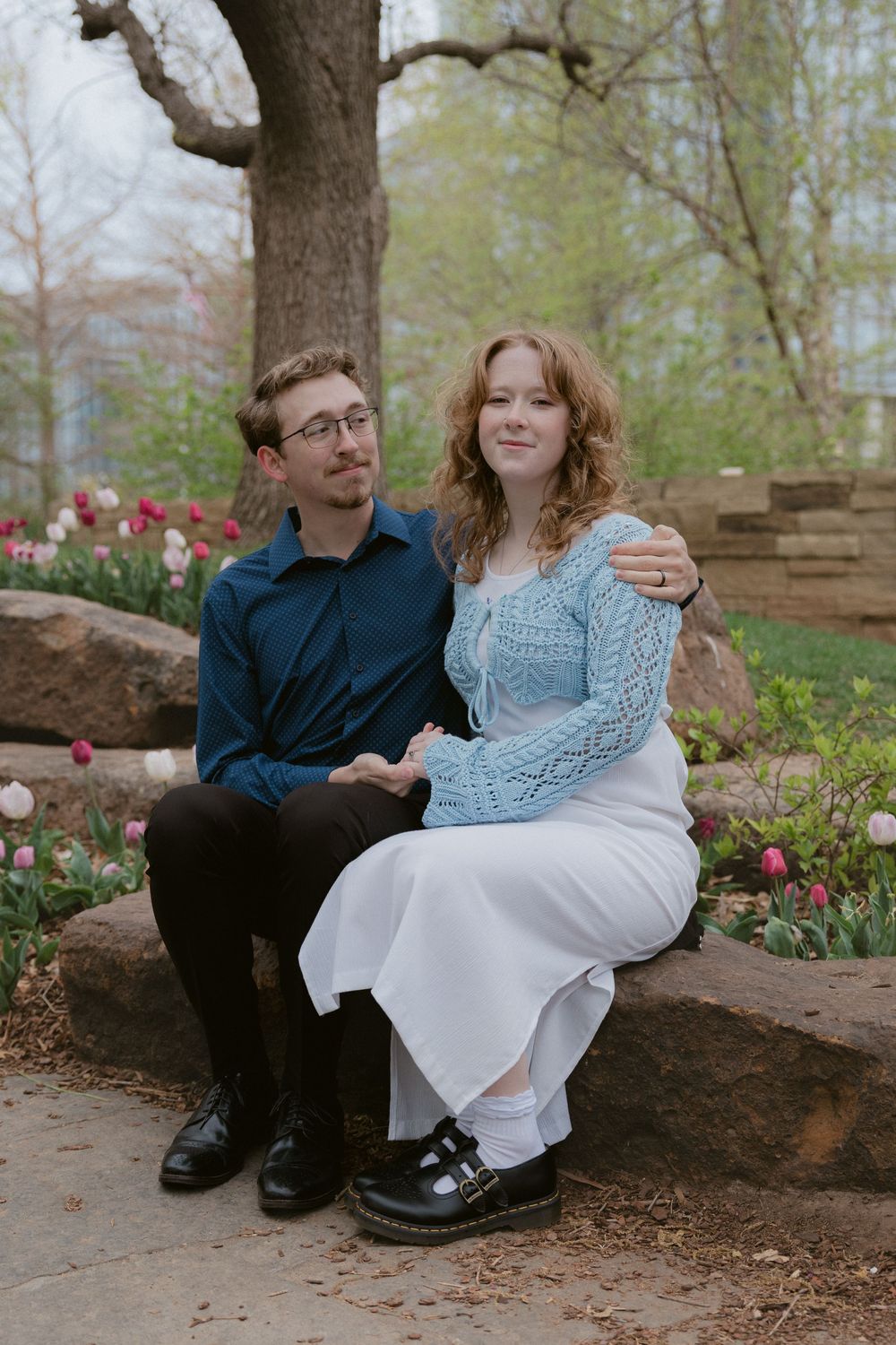 Couple sitting together on stone wall surrounded by spring flowers and trees.