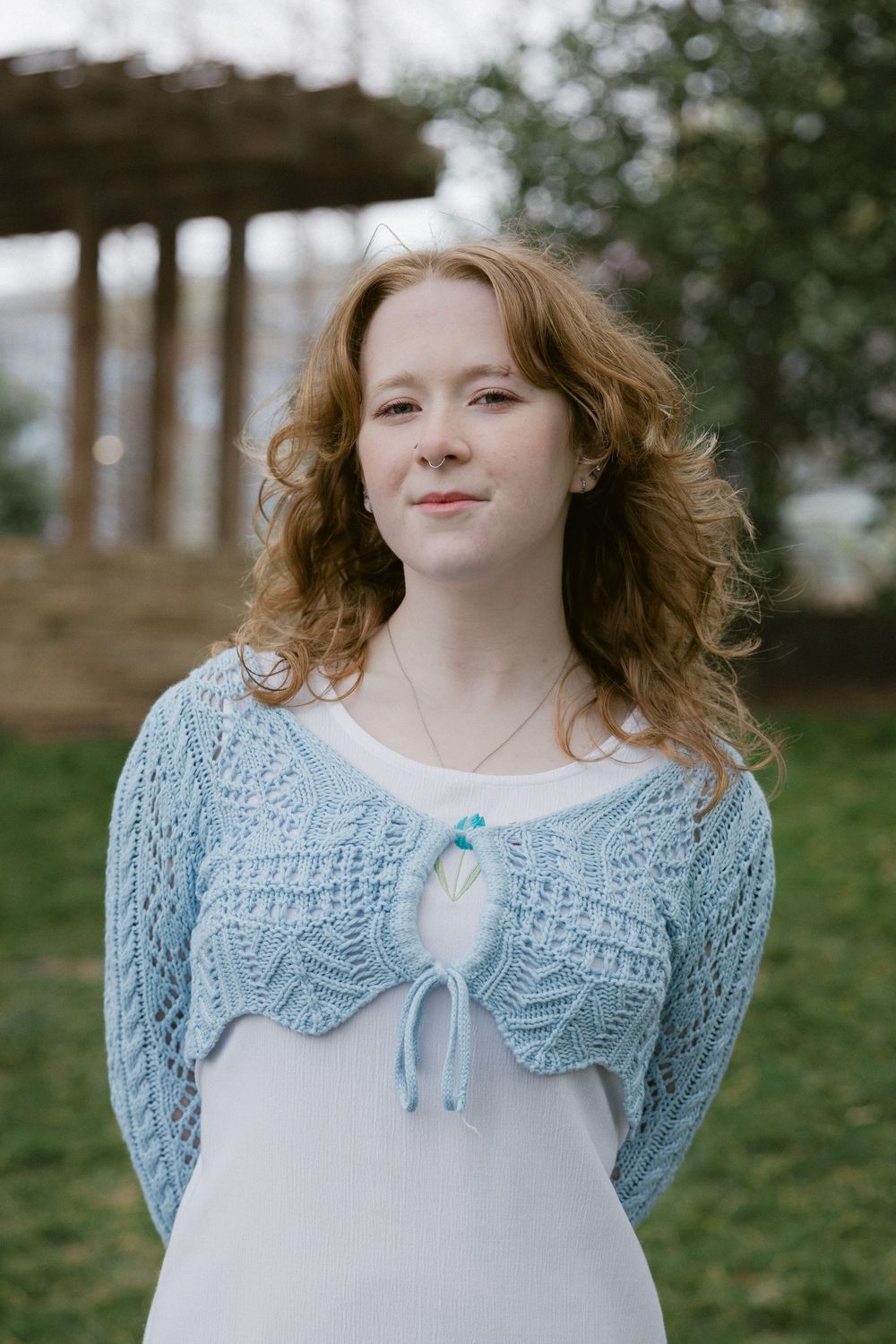 A person wearing a light blue knit cardigan and white dress standing in front of a rustic background.