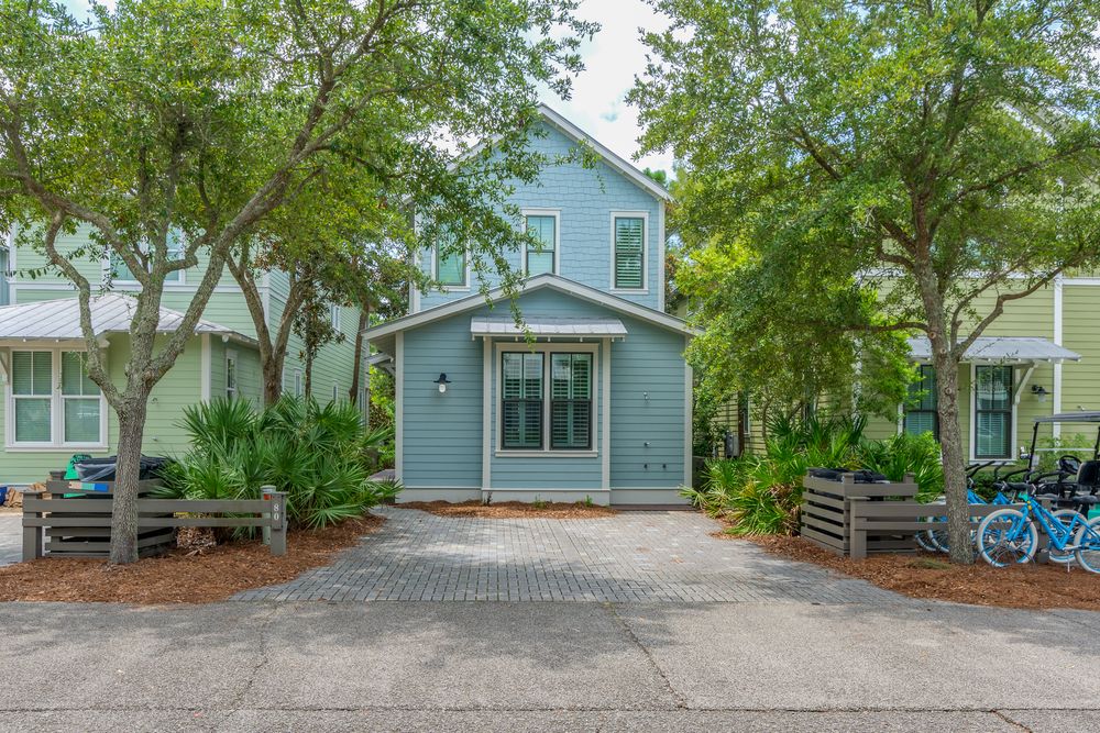 Four identical brick paved driveways lead to coastal style homes with blue siding surrounded by lush green trees.