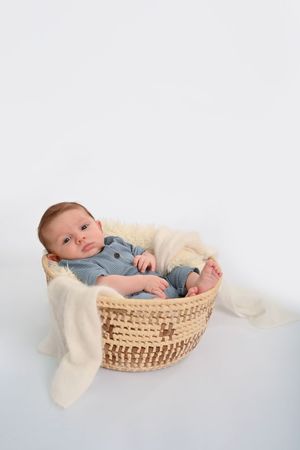 Baby wearing blue outfit resting in a woven basket with cream colored blanket.