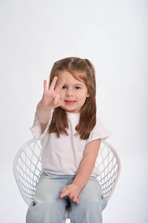 Child in white shirt making playful expressions while sitting in white chair.