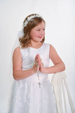 Person wearing white communion dress and veil standing beside white chair.