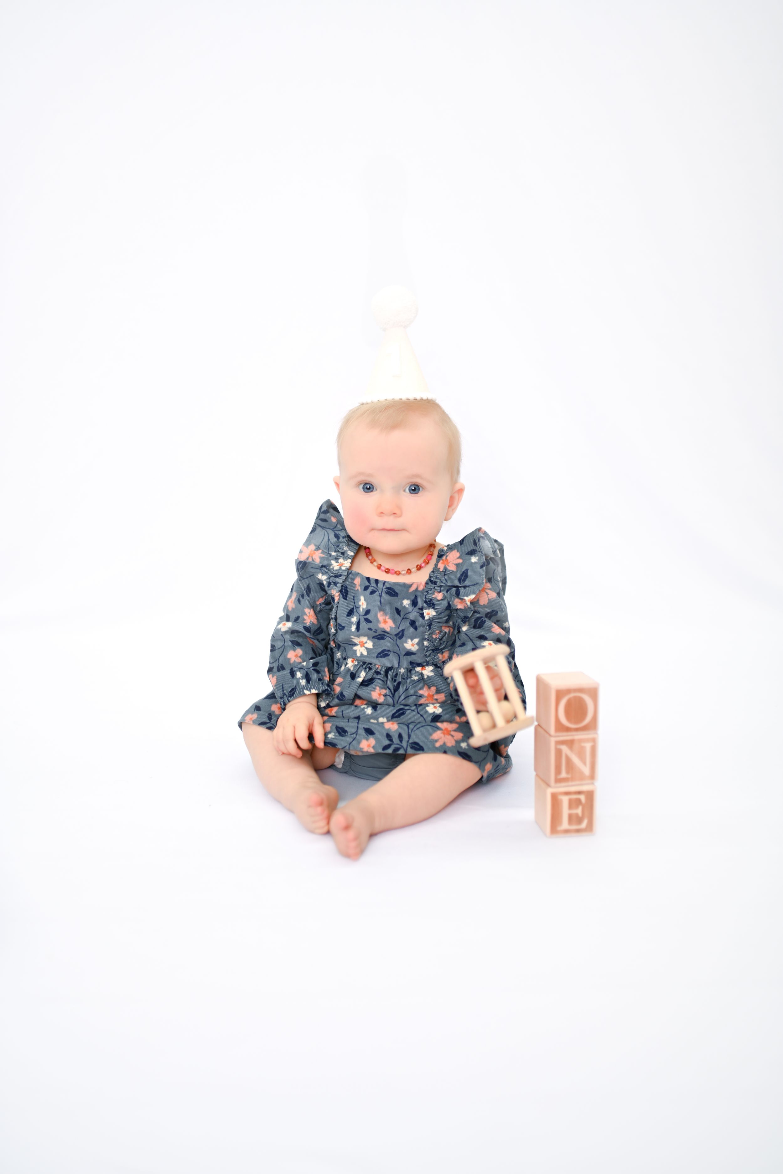 Baby in floral dress sits with wooden blocks spelling 'ONE' against white background.